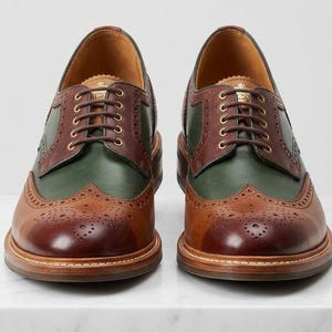 May include: A pair of leather brogue shoes featuring a multi-tone design with dark brown toe caps, forest green side panels, and medium brown lace-up sections. The shoes include decorative perforated detailing along the seams and brown laces.
