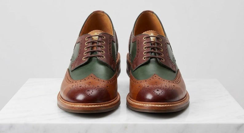 May include: A pair of leather brogue shoes featuring a multi-tone design with dark brown toe caps, forest green side panels, and medium brown lace-up sections. The shoes include decorative perforated detailing along the seams and brown laces.