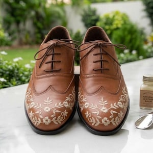 May include: A pair of tan leather oxford shoes featuring a wingtip design with intricate cream-colored floral embroidery on the toe caps. The shoes have dark brown laces, a low heel, and a smooth finish, showcasing detailed stitching throughout.