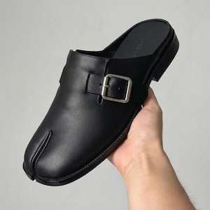 May include: A black leather mule shoe featuring a split-toe design, a low heel, and a wide strap across the top secured with a silver-toned metal buckle. The shoe has a smooth finish and an open back, showcasing a distinct cleft at the front toe area.