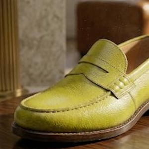 Puede incluir: Un solo mocas&iacute;n de cuero verde lima con una correa de centavo. El zapato tiene una suela marr&oacute;n y est&aacute; colocado sobre una superficie de madera oscura. El cuero tiene un aspecto texturizado y las costuras son visibles en los bordes.