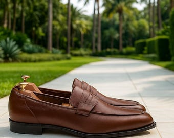 Può includere: Un paio di mocassini in pelle marrone dal design classico. Le scarpe hanno una consistenza liscia e una suola scura. Lo sfondo presenta un sentiero e una vegetazione lussureggiante, suggerendo un ambiente esterno. Le scarpe sono esposte con forme di legno.