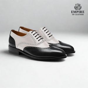 Puede incluir: Un par de zapatos Oxford de cuero bicolor en negro y blanco, con cordones negros y tac&oacute;n bajo. La secci&oacute;n blanca tiene perforaciones decorativas. El logotipo de Empire of Leather est&aacute; en la esquina superior derecha.