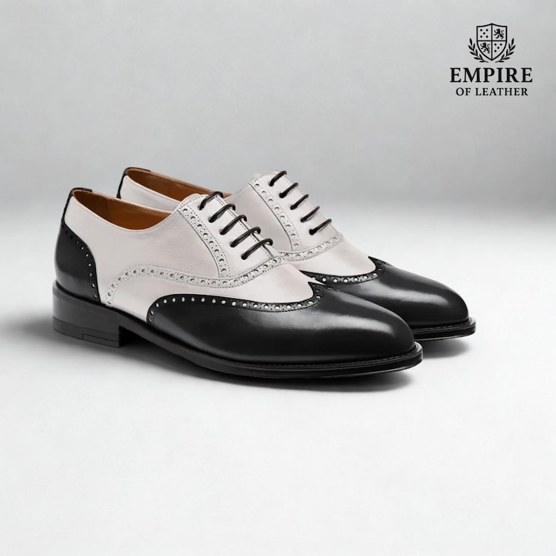 Puede incluir: Un par de zapatos Oxford de cuero bicolor en negro y blanco, con cordones negros y tac&oacute;n bajo. La secci&oacute;n blanca tiene perforaciones decorativas. El logotipo de Empire of Leather est&aacute; en la esquina superior derecha.