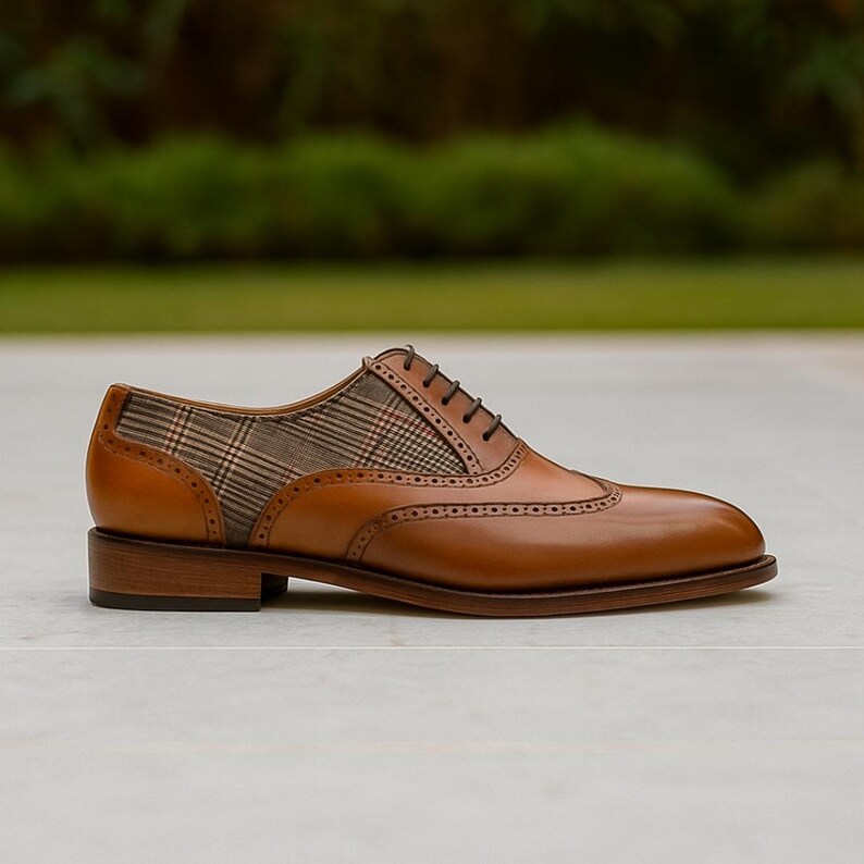 Op de afbeelding: Een bruine leren herenschoen met een geruit stoffen paneel. De schoen heeft een wingtip-ontwerp met decoratieve perforaties en donkerbruine veters. De schoen staat op een neutrale achtergrond.