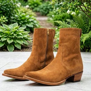 May include: A pair of tan suede ankle boots featuring a pointed toe, a stacked wooden heel, and a vertical gold-toned zipper closure on the inner side of each boot. The boots have a clean, minimalist silhouette with visible stitching along the vamp.