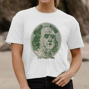 Puede incluir: Camiseta blanca con un retrato verde y beige de Benjamin Franklin, similar a un billete de cien dólares estadounidenses. La camiseta tiene mangas cortas y cuello redondo.