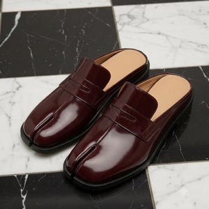 May include: A pair of dark burgundy patent leather mule loafers featuring a distinctive split-toe design. These slip-on shoes have a glossy finish, a traditional penny loafer strap across the vamp, and a beige interior lining with a flat sole.