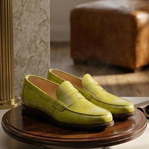 Puede incluir: Un par de mocasines de cuero amarillo brillante sobre un expositor de madera. Los zapatos tienen un dise&ntilde;o cl&aacute;sico con punta redondeada y una correa en el empeine. El expositor est&aacute; girando. El texto "HERITAGE COLLECTOR" es visible.