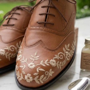 May include: A pair of tan leather oxford shoes featuring a wingtip design with intricate cream-colored floral embroidery on the toe caps. The shoes have brown laces, a low heel, and a dark sole, accompanied by a small glass bottle and a metal spoon.