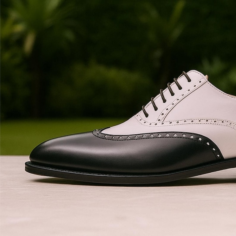 Puede incluir: Zapato Oxford de cuero de dos tonos. El zapato presenta una puntera y un tal&oacute;n negros, con una parte superior blanca. El zapato tiene cordones negros y perforaciones decorativas a lo largo de las costuras. El zapato est&aacute; sobre un fondo verde borroso.