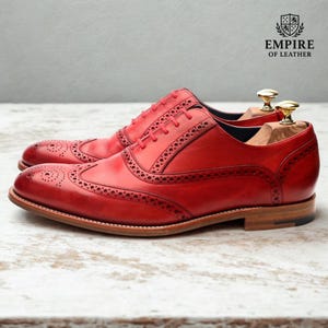 May include: A pair of red leather brogue shoes with intricate perforations and a classic wingtip design. The shoes have a light brown sole and are displayed with wooden shoe trees. The Empire of Leather logo is visible.