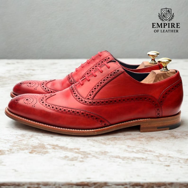 May include: A pair of red leather brogue shoes with intricate perforations and a classic wingtip design. The shoes have a light brown sole and are displayed with wooden shoe trees. The Empire of Leather logo is visible.