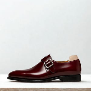 May include: A burgundy leather monk strap shoe with a silver buckle. The shoe has a black sole and a tan heel counter. The shoe is a classic style, suitable for formal occasions.