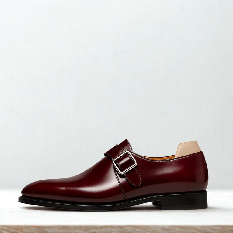 May include: A burgundy leather monk strap shoe with a silver buckle. The shoe has a black sole and a tan heel counter. The shoe is a classic style, suitable for formal occasions.