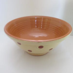 May include: A ceramic bowl with a cream-colored exterior and a reddish-orange interior. The bowl features evenly spaced maroon polka dots around the outside. The interior has a glossy finish.