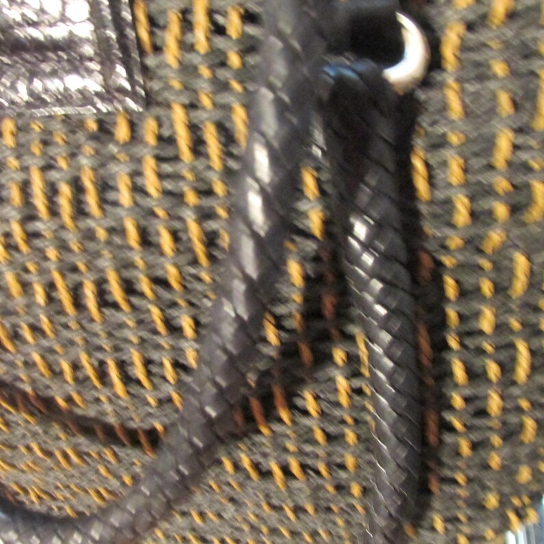 May include: Close-up of a woven handbag with a black leather strap and silver-tone hardware. The bag's weave features a pattern of black and gold threads, creating a textured surface. The strap is braided, adding to the bag's unique design.