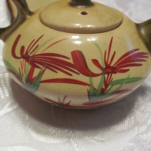 May include: A small, cream-colored teapot with a brown handle and lid. The teapot is decorated with red and green floral designs.