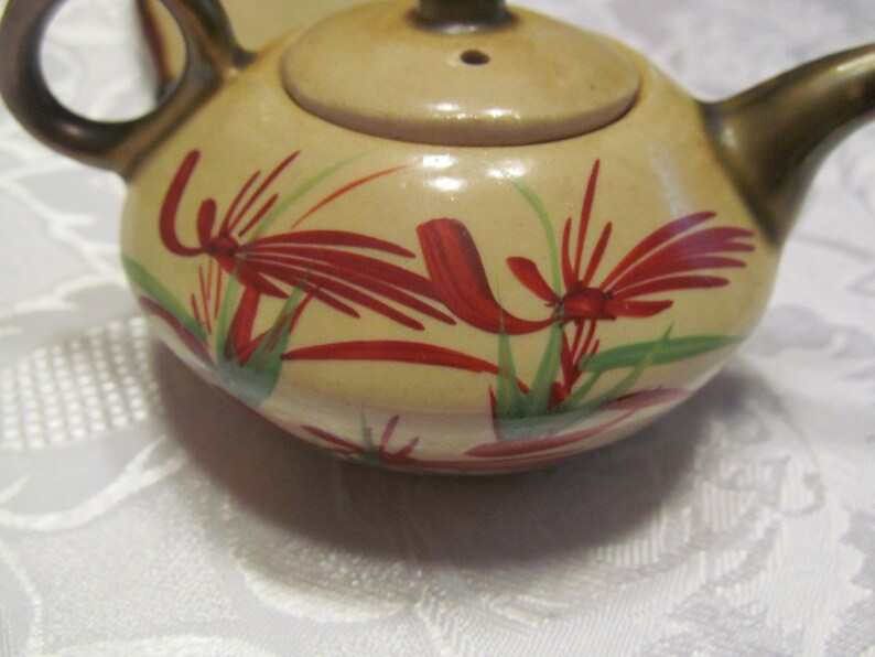 May include: A small, cream-colored teapot with a brown handle and lid. The teapot is decorated with red and green floral designs.