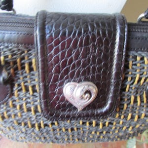 May include: Close-up of a brown and woven handbag with a flap closure. The flap is dark brown with a textured, crocodile-like pattern and a silver heart-shaped clasp. The bag's woven body features yellow and gray threads.