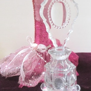 May include: A clear glass perfume bottle with a decorative stopper. The bottle has an octagonal shape with a rounded base. The stopper is a clear, ornate design with an oval center and a beaded border. A pink fabric with sequins is in the background.