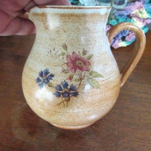 May include: A tan ceramic pitcher with a floral design featuring pink and blue flowers. The pitcher has a rounded body, a curved handle, and a spout. The pitcher is likely used for serving beverages.