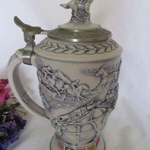 May include: A ceramic beer stein with a horse racing theme. The stein features a detailed relief of horses and riders in a race, with a horse and rider on top of the lid. The stein has a handle and a base with a colorful design.