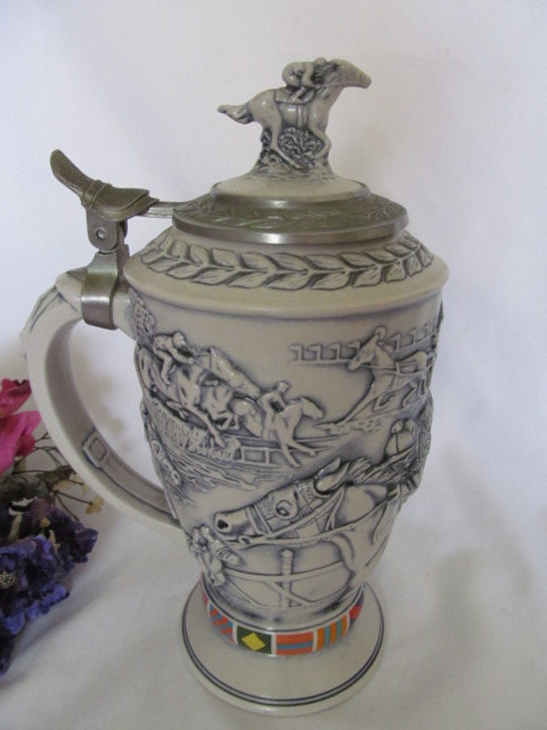 May include: A ceramic beer stein with a horse racing theme. The stein features a detailed relief of horses and riders in a race, with a horse and rider on top of the lid. The stein has a handle and a base with a colorful design.