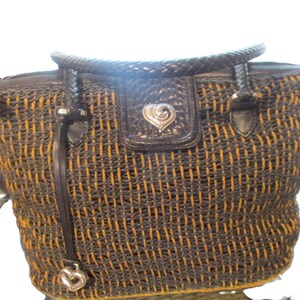 May include: A woven tote bag with a black and gold pattern. The bag has a braided handle and a black leather flap with a silver heart-shaped emblem. A silver heart charm hangs from a leather strap.
