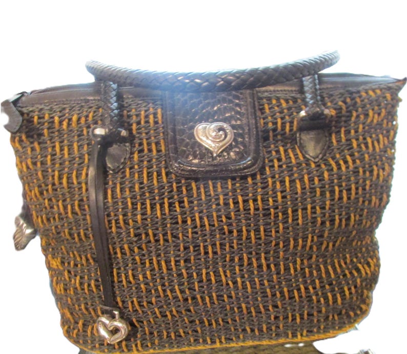 May include: A woven tote bag with a black and gold pattern. The bag has a braided handle and a black leather flap with a silver heart-shaped emblem. A silver heart charm hangs from a leather strap.