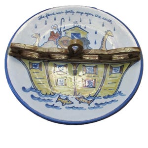 May include: A round ceramic plate featuring a hand-painted illustration of Noah's Ark on water with animals and a figure. The text "The flood was forty days upon the earth" is written at the top. A brown, textured, bridge-like handle spans the center.
