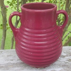 May include: A deep red ceramic vase with two handles and a ribbed design. The vase is sitting on a wooden surface.