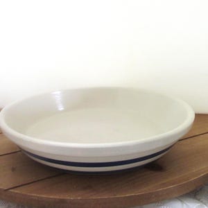 May include: A white ceramic pie dish with two navy blue stripes around the exterior. The dish is round with a slightly raised rim. It sits on a wooden surface.