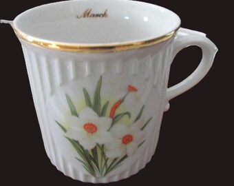 May include: A white porcelain mug featuring a vertical ribbed texture and a gold-trimmed rim. The exterior displays a floral illustration of white daffodils with orange centers. The interior rim is inscribed with the word March in gold script.