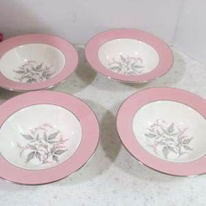 May include: A set of four round ceramic bowls featuring a wide pink rim and a silver-toned edge. The center of each bowl is off-white and decorated with a botanical illustration of grey leaves and small pink flowers arranged in a central cluster.