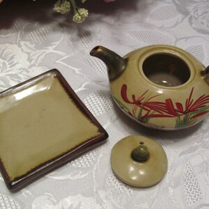 May include: A small, brown ceramic teapot with a floral design in red, green, and white. The teapot has a lid and a matching square saucer.