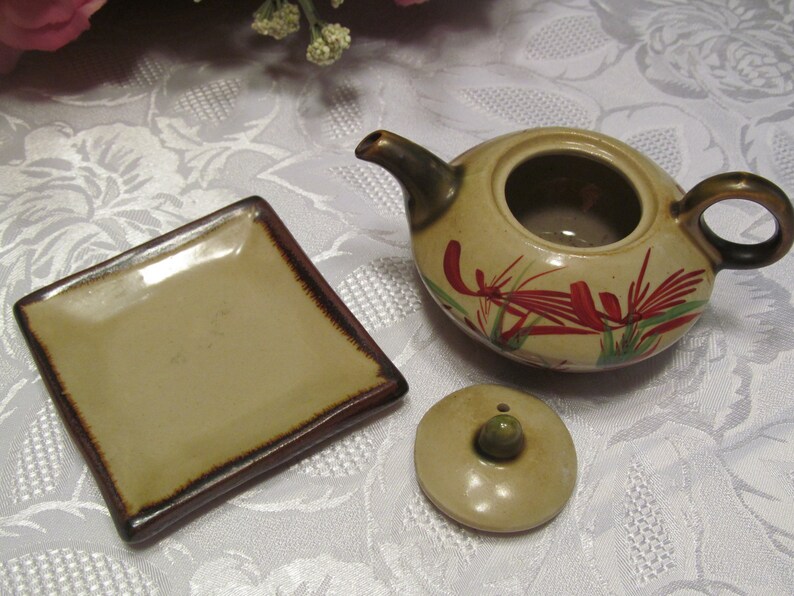 May include: A small, brown ceramic teapot with a floral design in red, green, and white. The teapot has a lid and a matching square saucer.