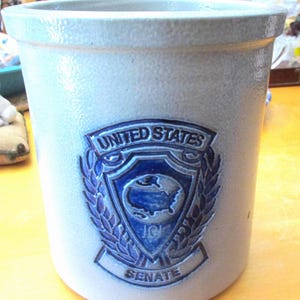 May include: A stoneware crock with a light gray glaze. A blue and white emblem of the United States Senate is centered on the front. The emblem features a shield with a map of the United States.