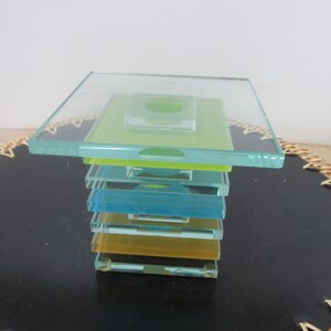 May include: A modern glass sculpture composed of stacked, rectangular glass plates in shades of clear, green, blue, and yellow. The top layer is a large, clear square. The sculpture has a minimalist design.