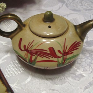 May include: A small, cream-colored teapot with a brown handle and spout. The teapot has a floral design with red and green flowers.