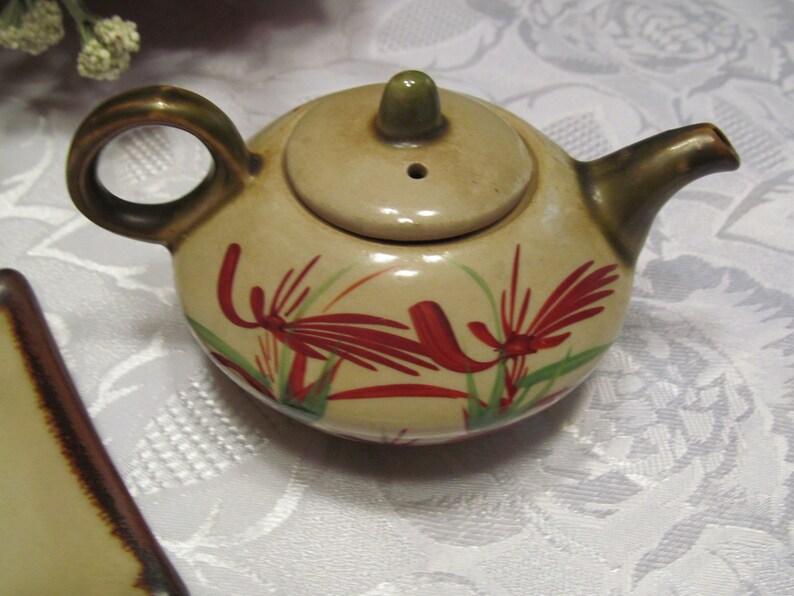 May include: A small, cream-colored teapot with a brown handle and spout. The teapot has a floral design with red and green flowers.