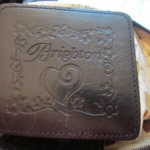 May include: Dark brown leather wallet with the embossed word "Brighton" surrounded by floral and heart designs. The wallet has a rectangular shape and a stitched border.