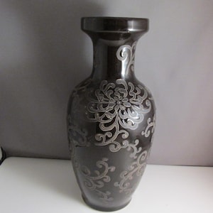May include: A dark brown ceramic vase with a silver floral design. The vase has a wide base and a narrow neck.