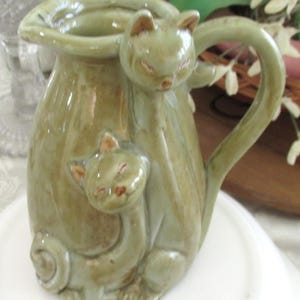 May include: A sage green ceramic pitcher featuring two sculpted cat figures. One cat stands tall forming part of the handle, while the second smaller cat sits at the base. The pitcher has a glossy, mottled glaze finish and a wide pouring spout.