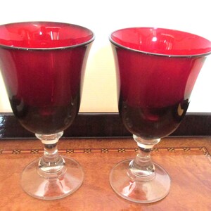 May include: Two matching wine glasses featuring deep ruby red bowls that transition into clear glass stems and circular bases. The glasses have a smooth, curved silhouette with a wide rim and a solid, transparent foot for stability on flat surfaces.