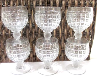 May include: Six clear, stemmed wine glasses. Each glass features a round bowl with a grid pattern, a small decorative ball, and a circular base. The glasses are arranged in front of a woven brown background.