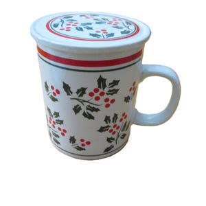 May include: White ceramic mug with a matching lid, adorned with a festive holly and berry pattern. The mug features a red stripe and green accents, perfect for holiday beverages. Includes a curved handle.
