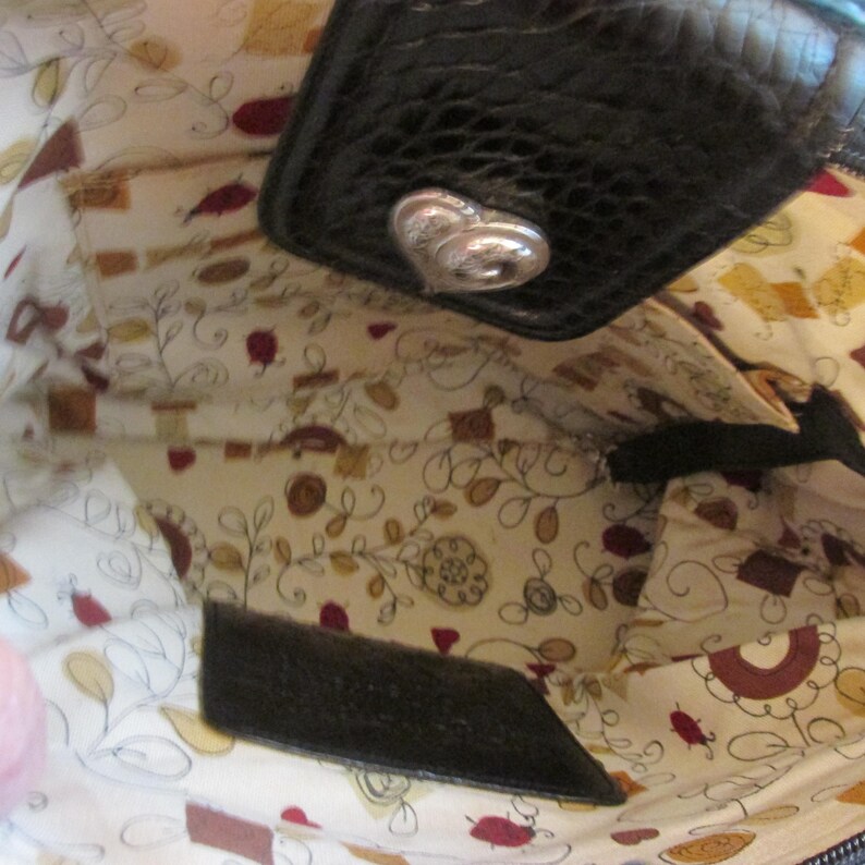 May include: Interior view of a black leather handbag with a silver heart-shaped clasp. The lining features a cream-colored fabric with a floral and leaf pattern in shades of brown, red, and yellow. A small pocket is visible.