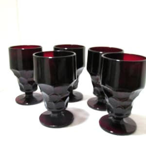 May include: A set of five deep red glass goblets featuring a faceted geometric pattern on the lower bowl and a smooth upper rim. Each goblet stands on a circular base, showcasing a consistent dark ruby color throughout the translucent glass material.