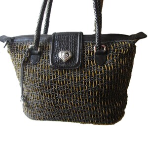 May include: Woven tote bag with black and gold threads. Features a black leather flap with a silver heart-shaped clasp. The bag has braided black handles and a zippered top closure.